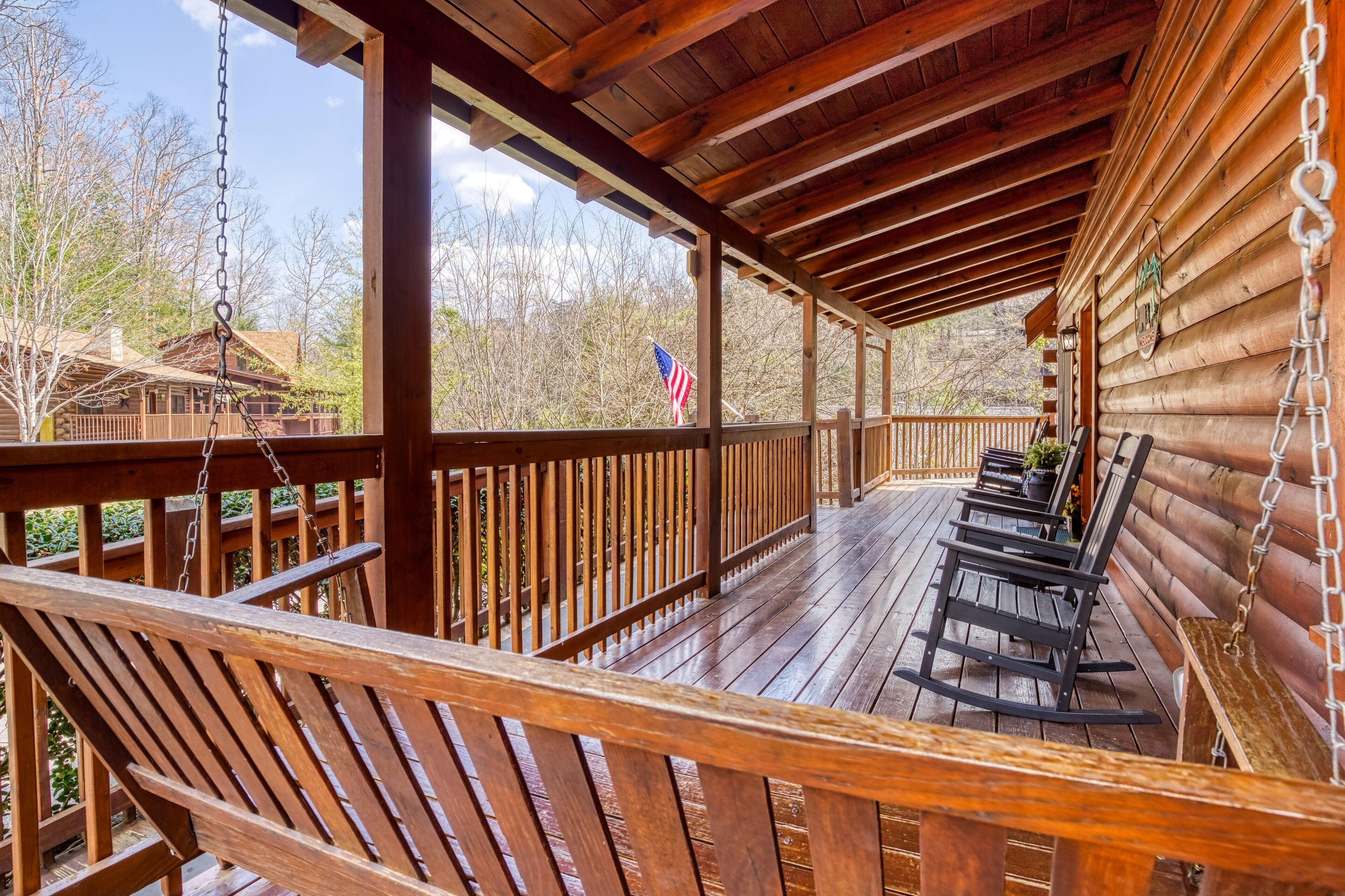 A wooden deck with rocking chairs, a flag, and a view of a cabin.