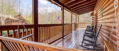 A wooden deck with rocking chairs, a flag, and a view of a cabin.