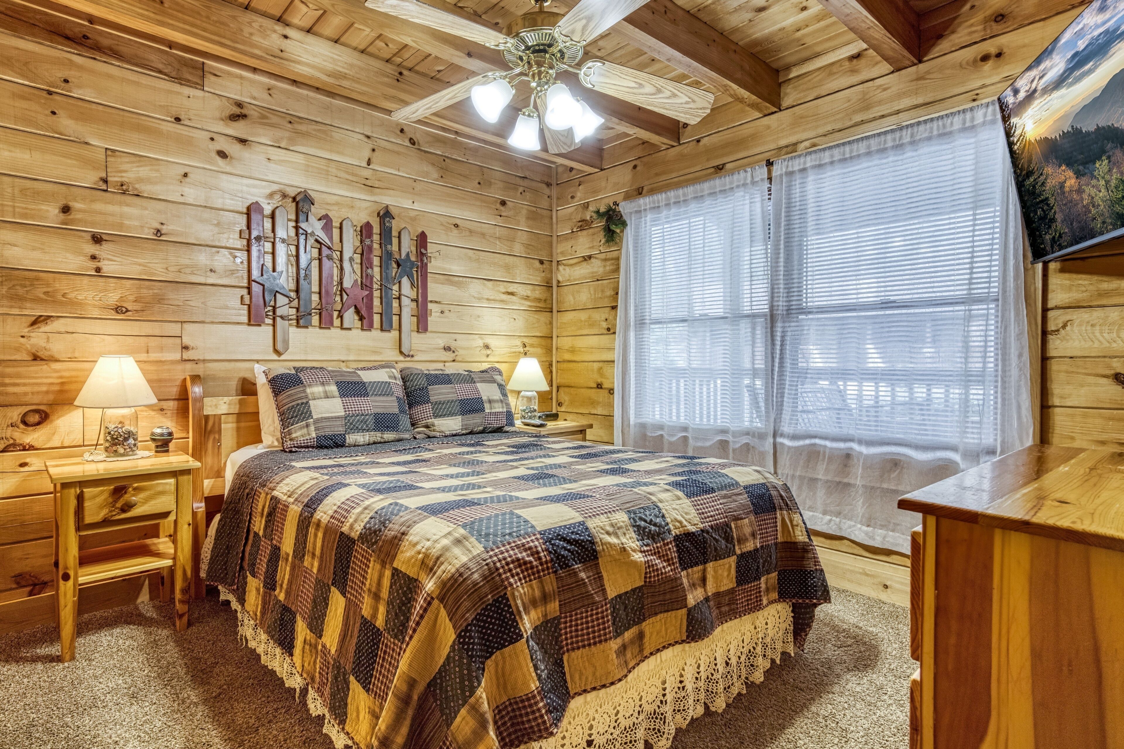 A wooden cabin bedroom with a bed, two bedside tables with lamps, a ceiling fan, and a window with sheer curtains.