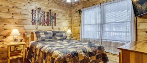 A wooden cabin bedroom with a bed, two bedside tables with lamps, a ceiling fan, and a window with sheer curtains.