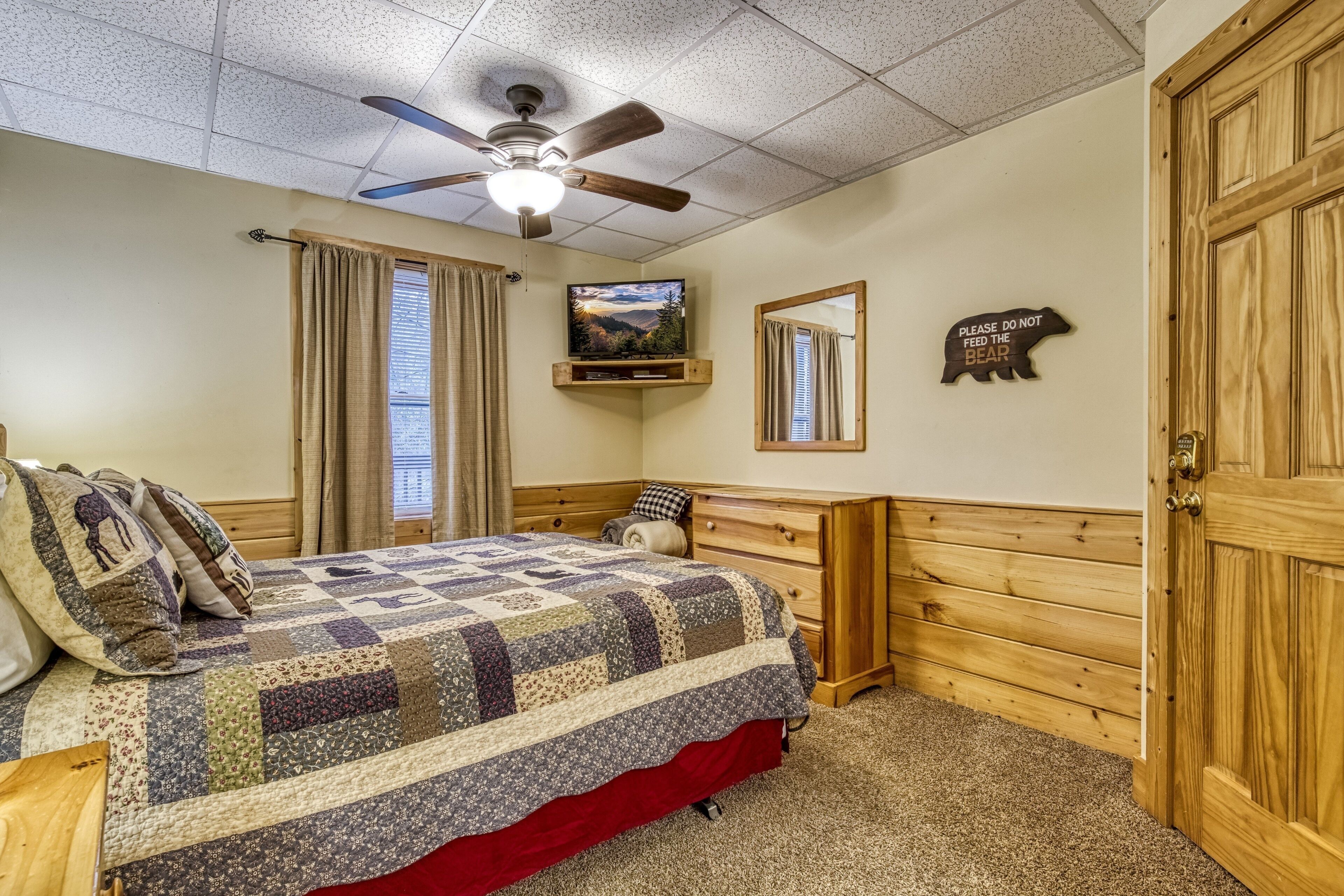 A bedroom with a wooden headboard, a quilted bedspread, a ceiling fan, a television on a shelf, a mirror, and a door with a bear sign.