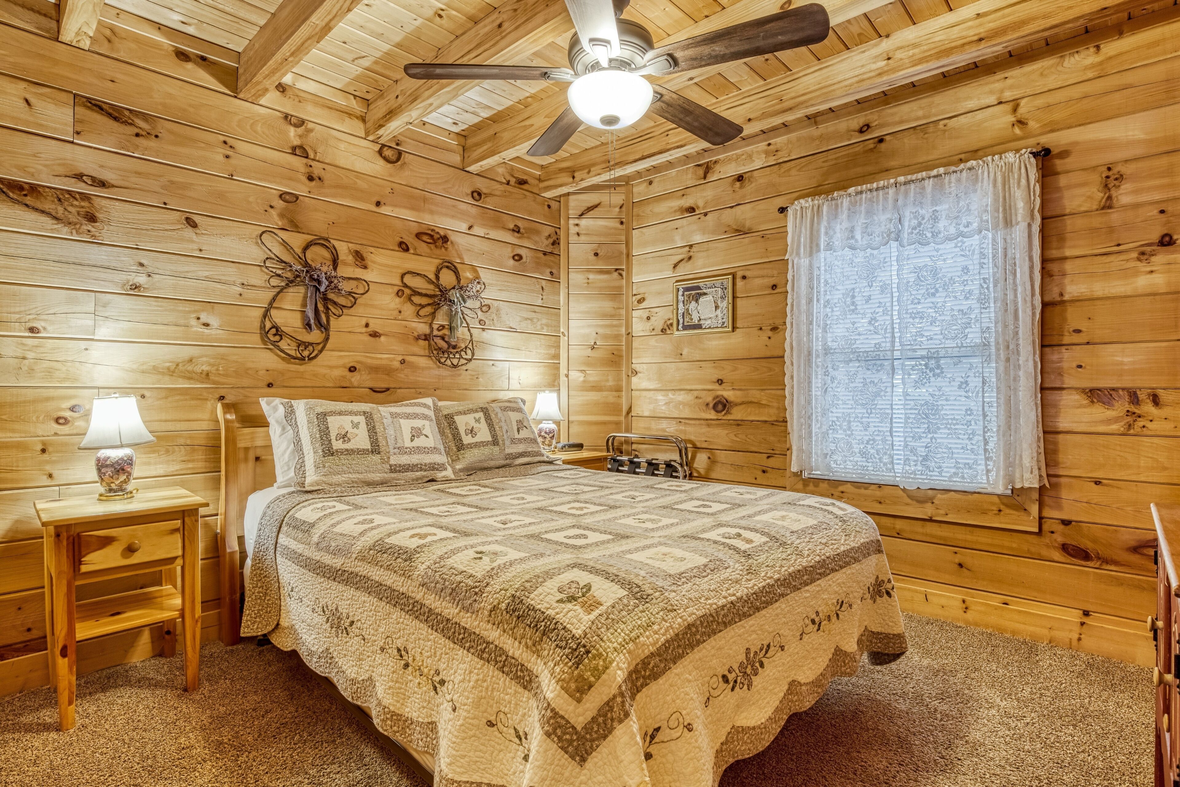 A wooden cabin bedroom with a bed, a ceiling fan, a window with curtains, and a small table with a lamp.