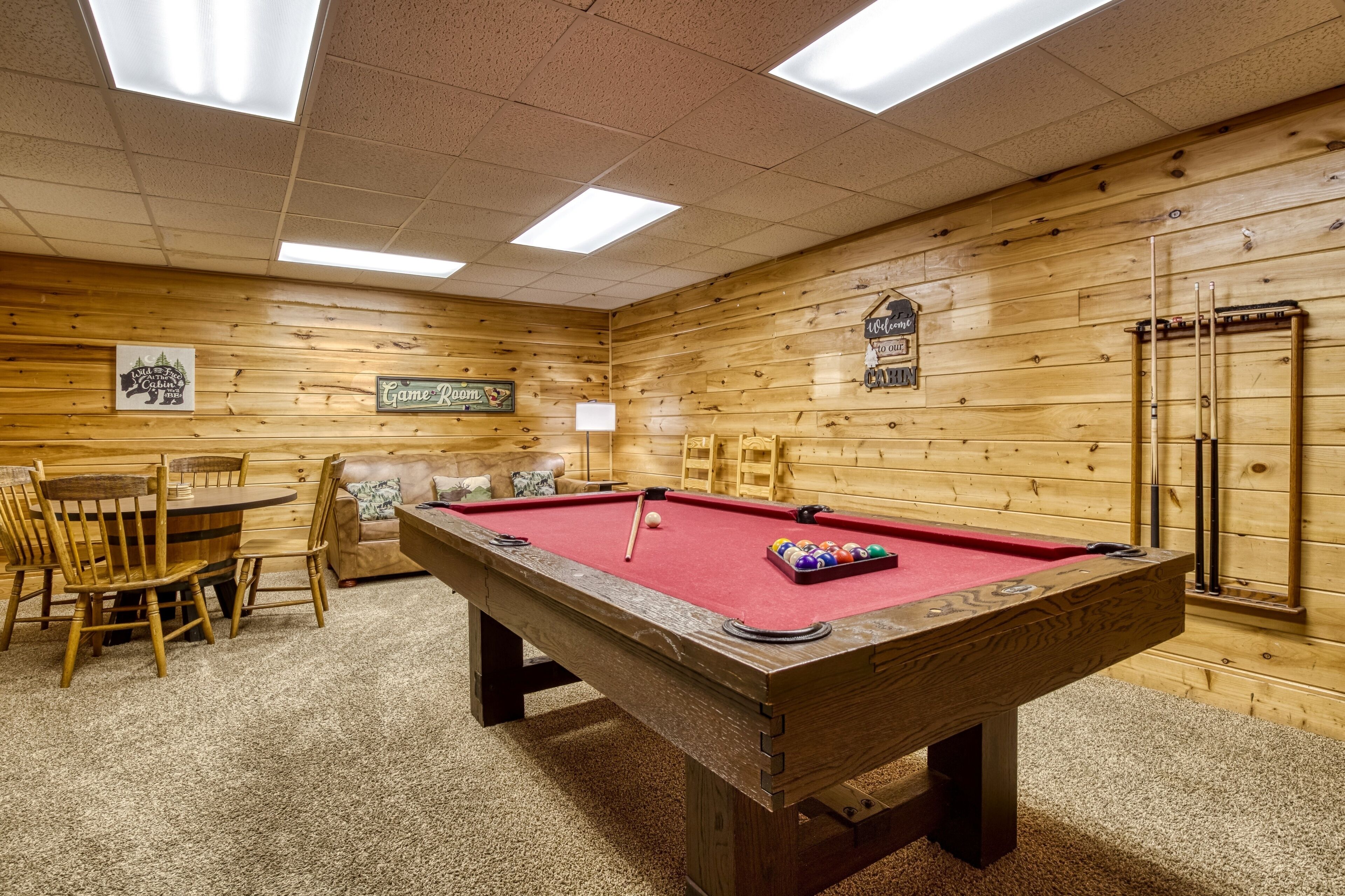A pool table with a red felt surface, surrounded by wooden chairs and a small table.