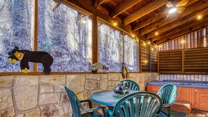 Una cabaña de madera con paredes de piedra, una mesa de piedra con sillas, un jacuzzi y una decoración de oso en el alféizar de la ventana.