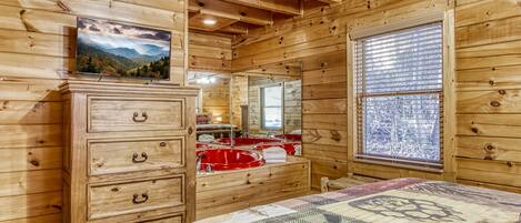 A wooden cabin room with a bed, a dresser, a mirror, and a window with blinds.