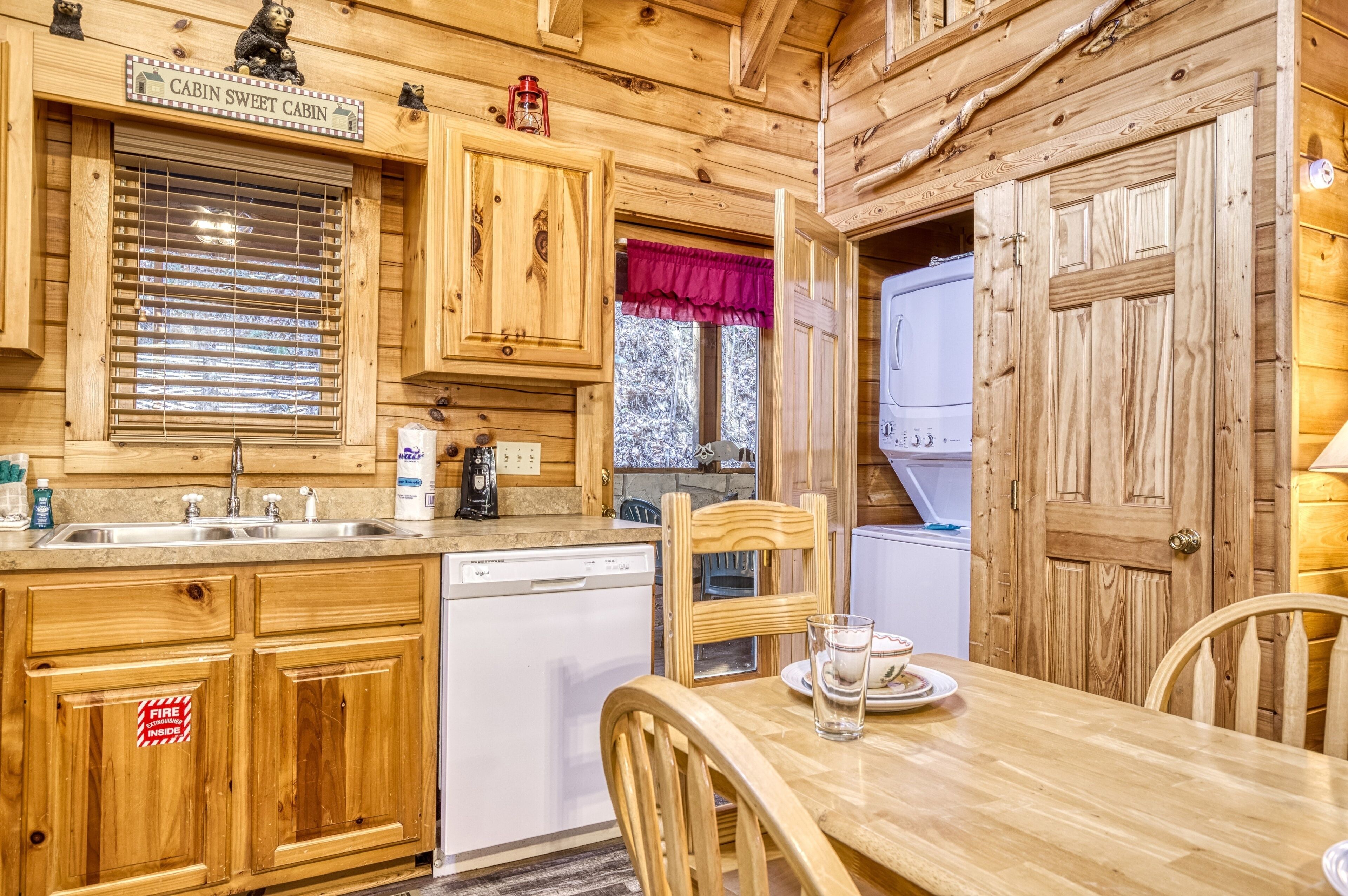 A cabin kitchen with wooden cabinets, a dishwasher, a dining table with chairs, and a window with a red valance.