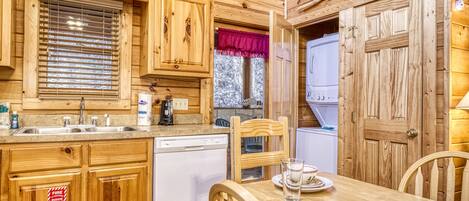 A cabin kitchen with wooden cabinets, a dishwasher, a dining table with chairs, and a window with a red valance.