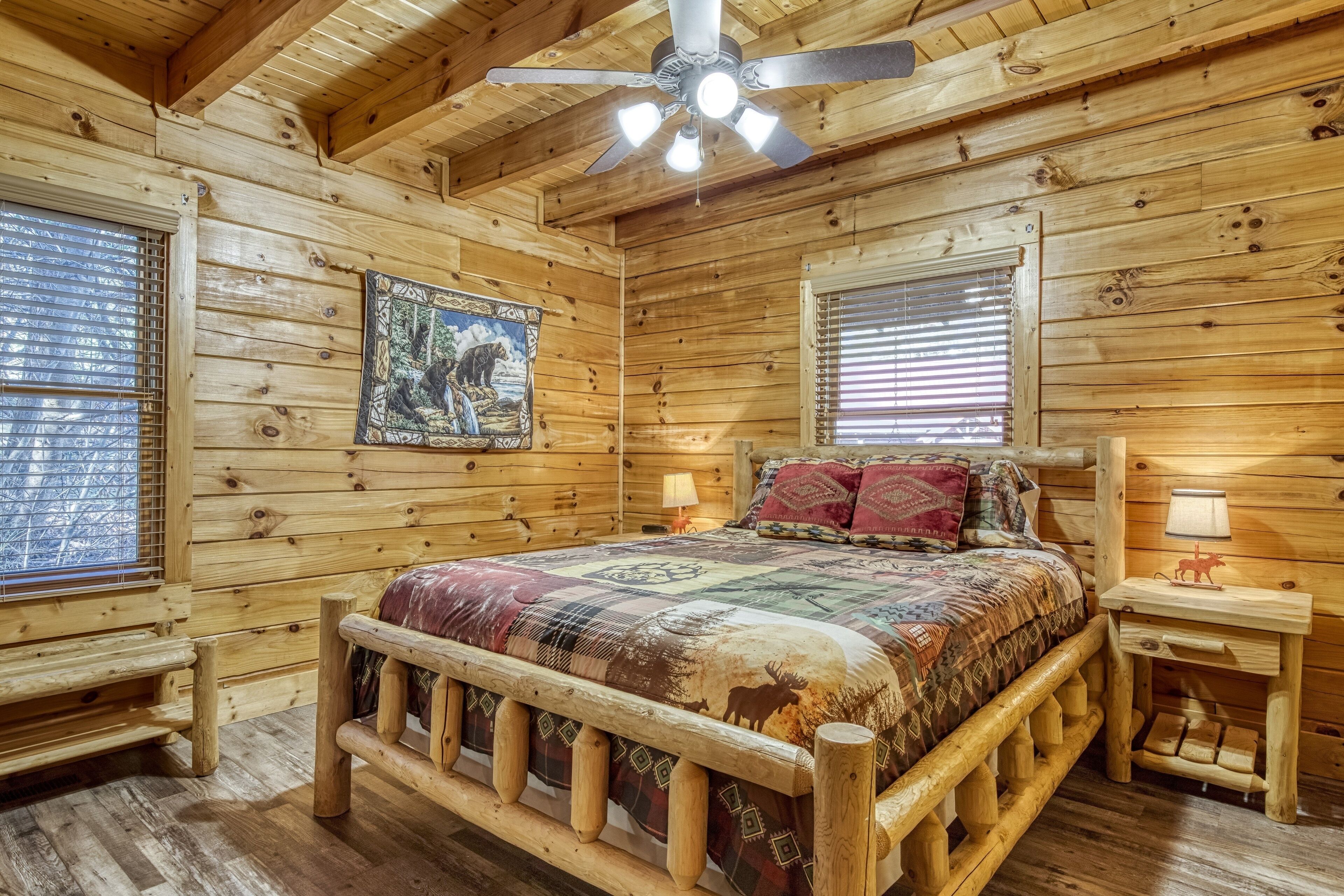 Un dormitorio de cabaña de madera con una cama, dos mesitas de noche, un ventilador de techo y ventanas con persianas.