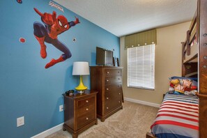 Une chambre avec un autocollant de Spiderman sur le mur, une commode, une lampe, un téléviseur posé sur un meuble, une fenêtre avec des stores et un lit avec un oreiller à l’effigie d’un super-héros.