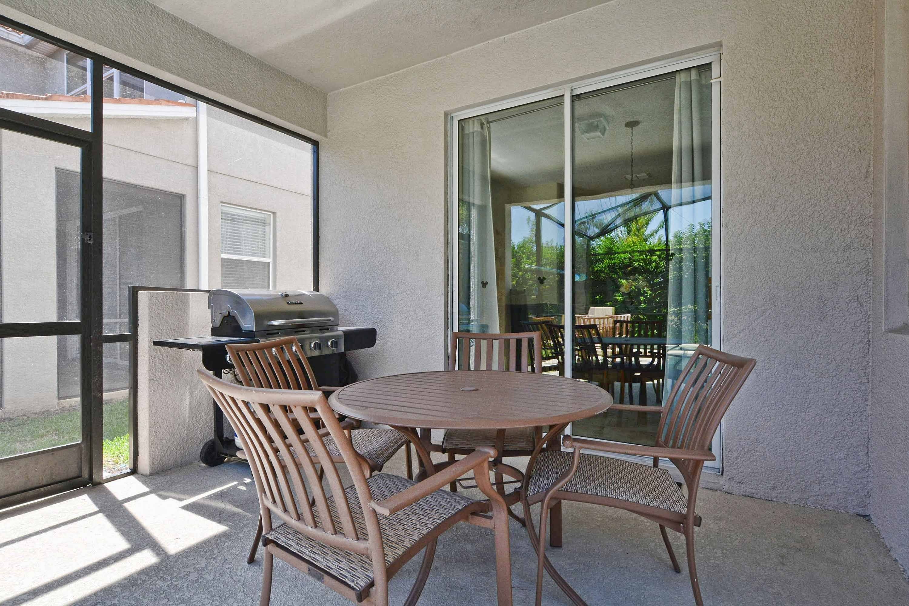 An outdoor patio with a round table and four chairs, a grill, and a sliding glass door leading to a patio area with outdoor furniture.
