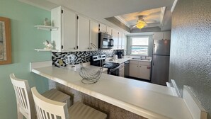 A kitchen with white cabinets, a black and white backsplash, a microwave, a stove, and a refrigerator. There is a window with a view of the beach. - 451 El Matador 2 Bedroom Condo by RedAwning (Fort Walton Beach)