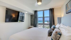 A hotel room with a bed, a TV, a window with a view of the beach, and a ceiling light.