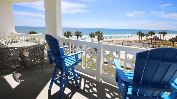 A balcony with blue chairs and a table, overlooking a beach with palm trees and umbrellas.