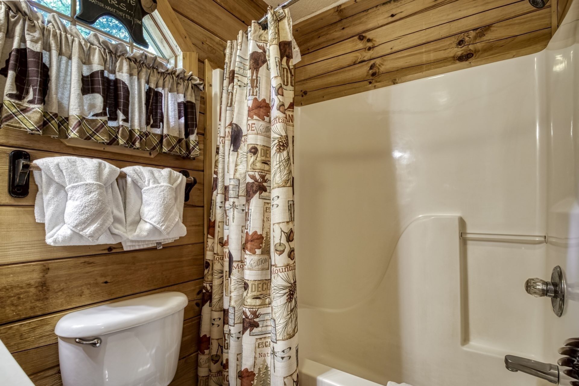 Een badkamer met houten inrichting, een toilet, een douchegordijn met natuurprint en handdoeken aan een rek.