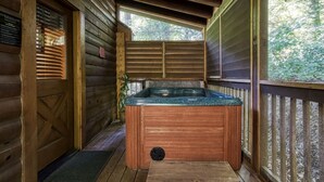 A wooden cabin with a hot tub on a wooden deck, surrounded by trees.
