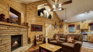 A cozy cabin interior with a stone fireplace, wooden walls, a sofa set, and a kitchen area.