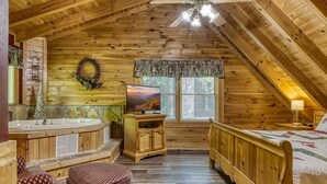 A cozy attic bedroom with a wooden ceiling, a television, a window with curtains, a bed, and a small seating area. - Er215 - Red Bird Haven Great Location - Close To Town! 3 Bedroom Cabin by RedAwning (Pigeon Forge)
