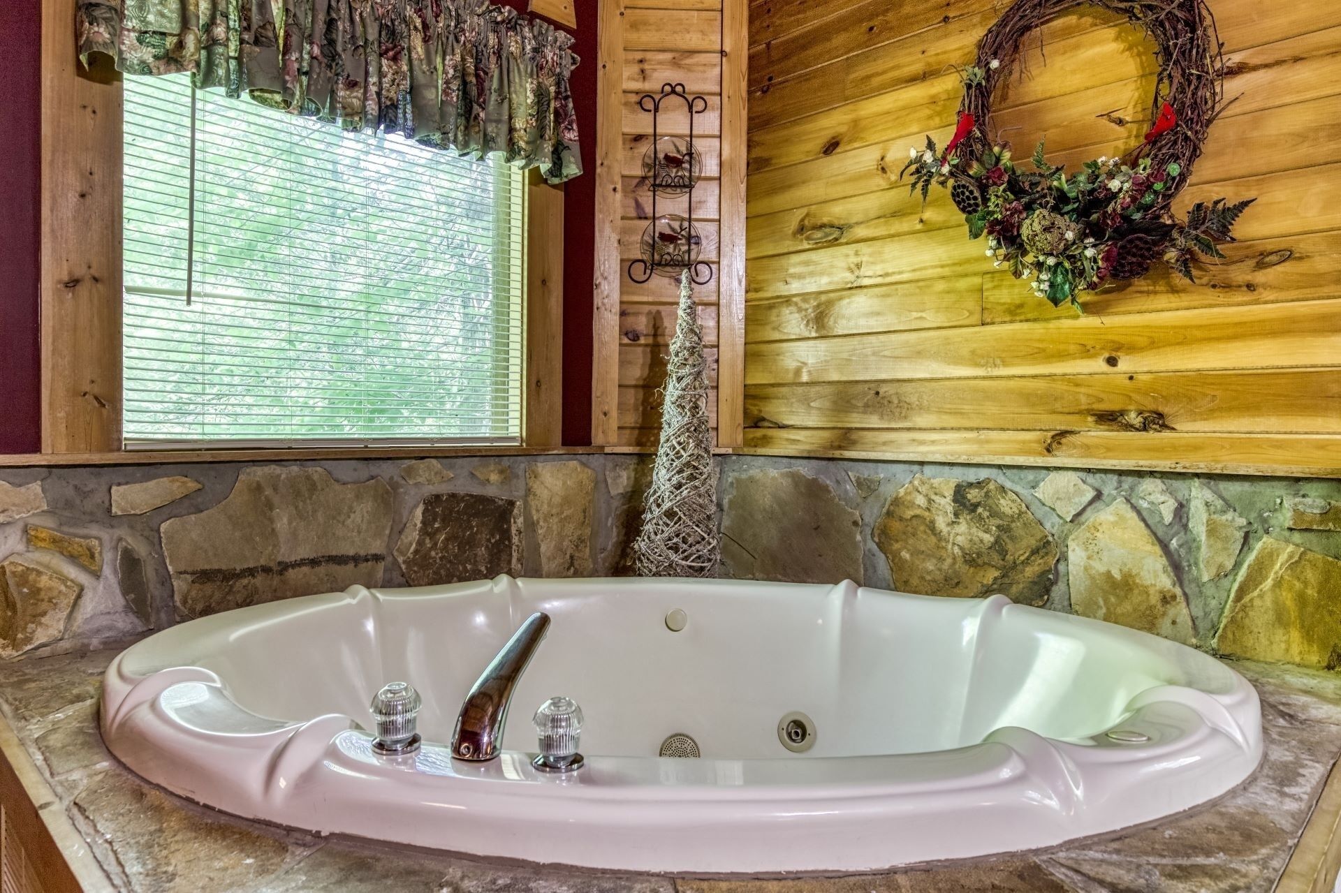 Un baño con pared de piedra, ventana de madera con persianas, bañera blanca y corona de madera en la pared.