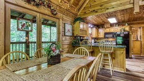A wooden cabin interior with a dining area, kitchen, and large windows. - Er215 - Red Bird Haven Great Location - Close To Town! 3 Bedroom Cabin by RedAwning (Pigeon Forge)