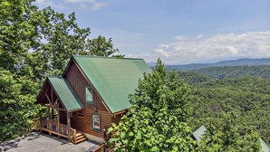 A rustic cabin with a green roof, wooden exterior, and a porch. It is surrounded by lush greenery and overlooks a forested valley. - Majestic Mountain View - Great Location! - Convenient To Everything! 2 Bedroom Cabin by RedAwning (Pigeon Forge)