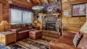 A cozy cabin interior with a stone fireplace, leather furniture, and a large window with blinds. - Majestic Mountain View - Great Location! - Convenient To Everything! 2 Bedroom Cabin by RedAwning (Pigeon Forge)
