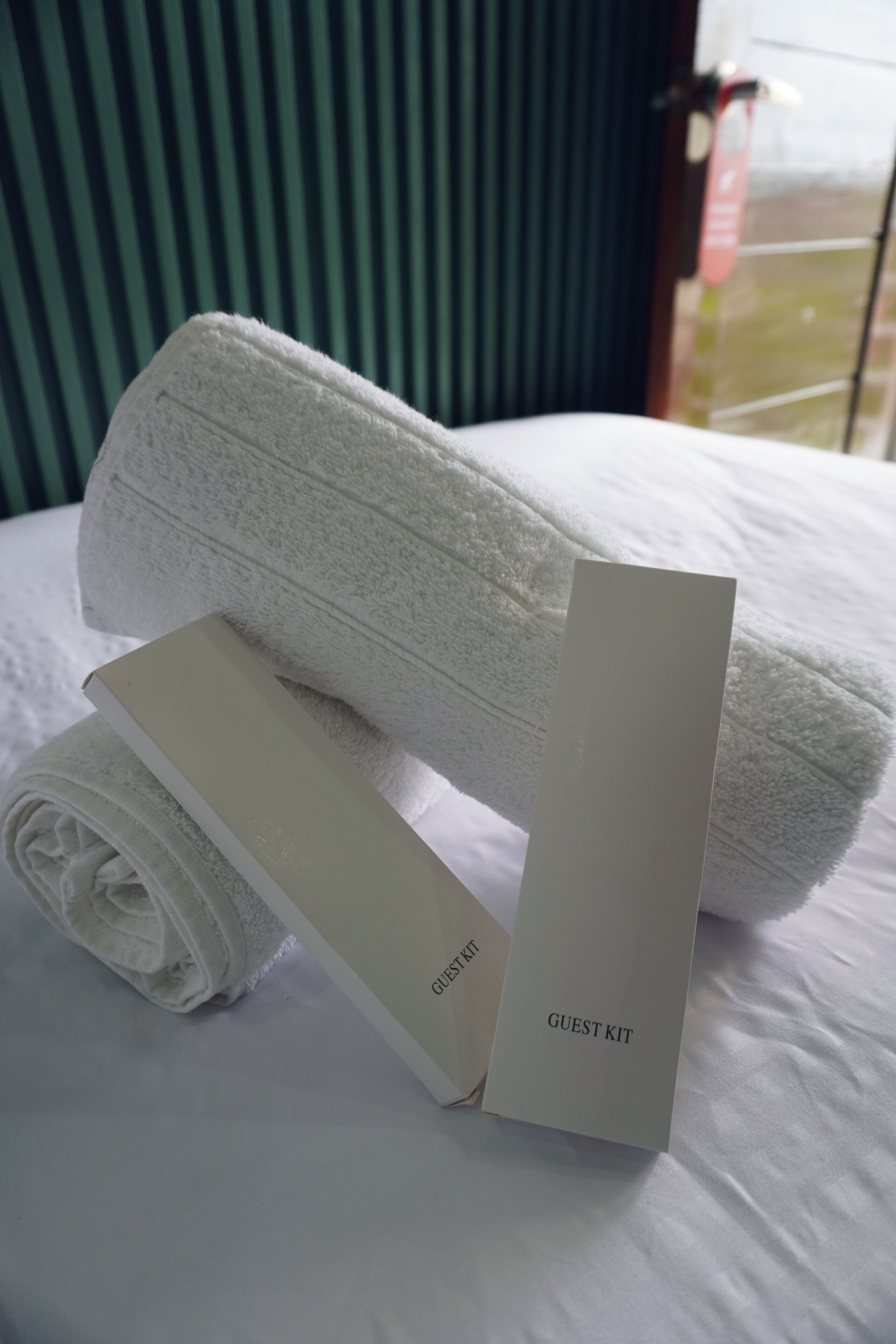 Una toalla blanca doblada con cuidado y una caja blanca de artículos de cortesía sobre una cama con ropa de cama blanca.