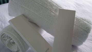 Una toalla blanca doblada con cuidado y una caja blanca de artículos de cortesía sobre una cama con ropa de cama blanca.