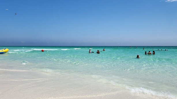 Ein Strand mit klarem türkisblauem Wasser, Menschen beim Schwimmen und Spielen, und ein gelbes Luftmatratze.