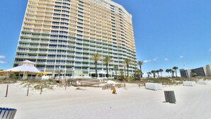 Un hôtel de grande hauteur doté de nombreux balcons, une plage bordée de palmiers et un ciel d’un bleu éclatant.