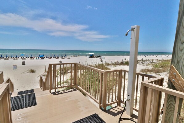 Een houten terras met een metalen leuning, dat uitkomt op een zandstrand met parasols en strandstoelen.