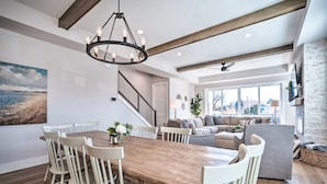 A spacious living area with a wooden dining table, white chairs, a gray sofa, and a large window offering a view of the city.