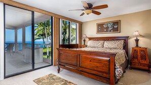 A bedroom with a large bed, a ceiling fan, a nightstand, and a view of the ocean through a sliding glass door.