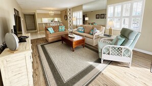 A spacious living room with a large sofa, a coffee table, and a chair. There is a kitchen area in the background, and a dining table with chairs.