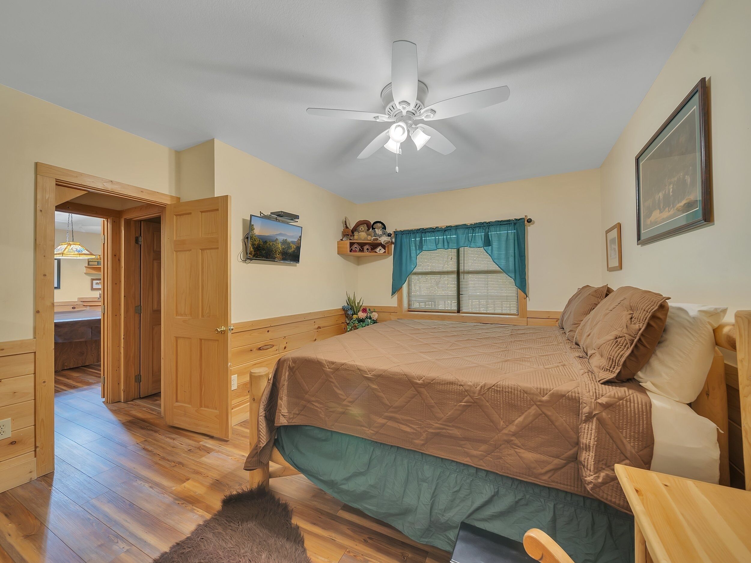 Un dormitorio con cama, piso de madera, ventilador de techo, ventana con cortinas y televisor en la pared.