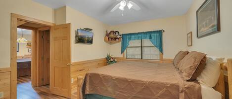 Un dormitorio con cama, piso de madera, ventilador de techo, ventana con cortinas y televisor en la pared.