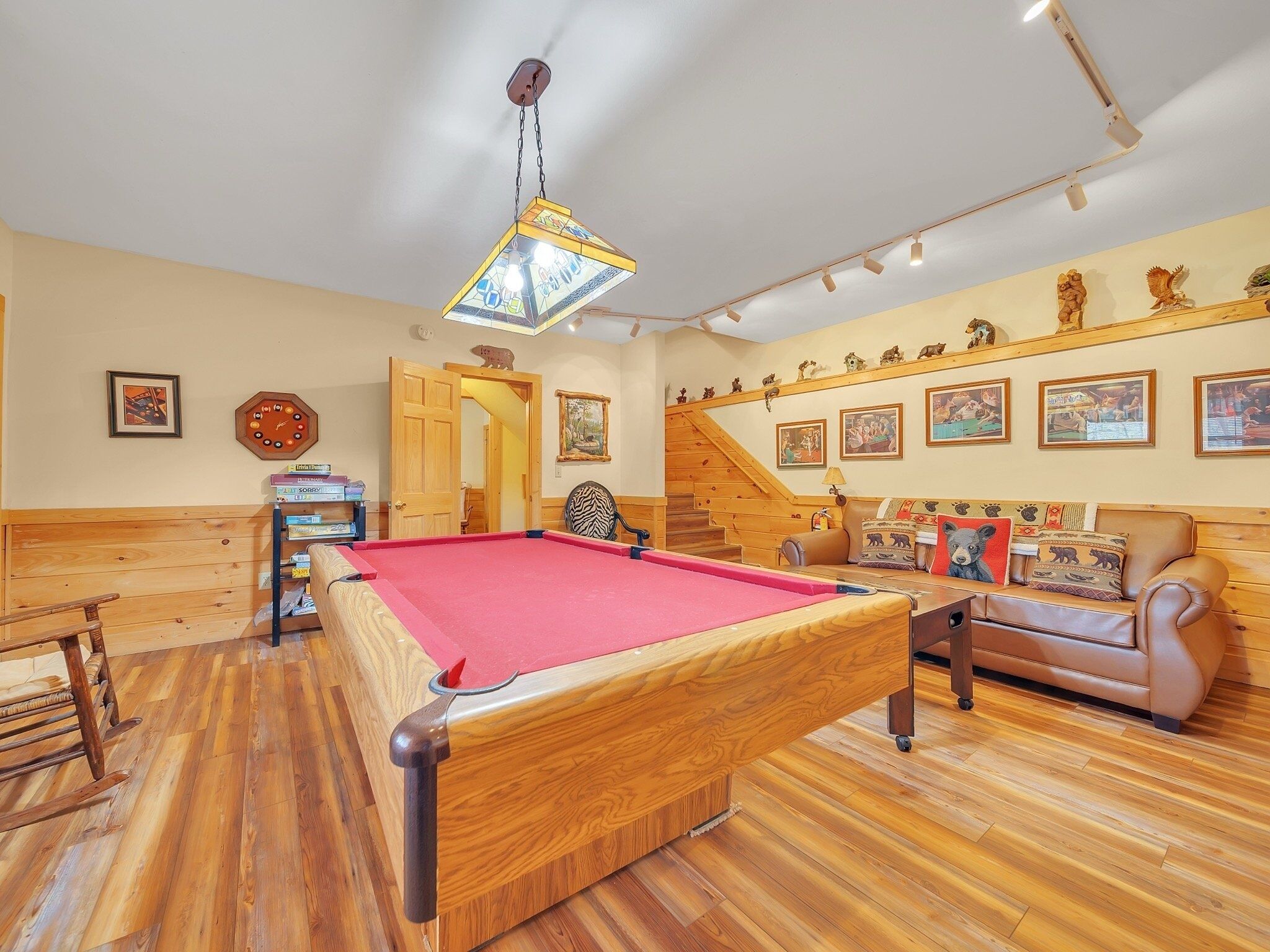 A billiards table in a room with wooden flooring, a wooden bench, a sofa, and a staircase.