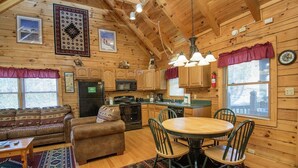Interior acogedor de una cabaña con detalles de madera, un comedor con una mesa redonda y sillas, una cocina con armarios de madera, y una sala de estar con un sofá y una chimenea.