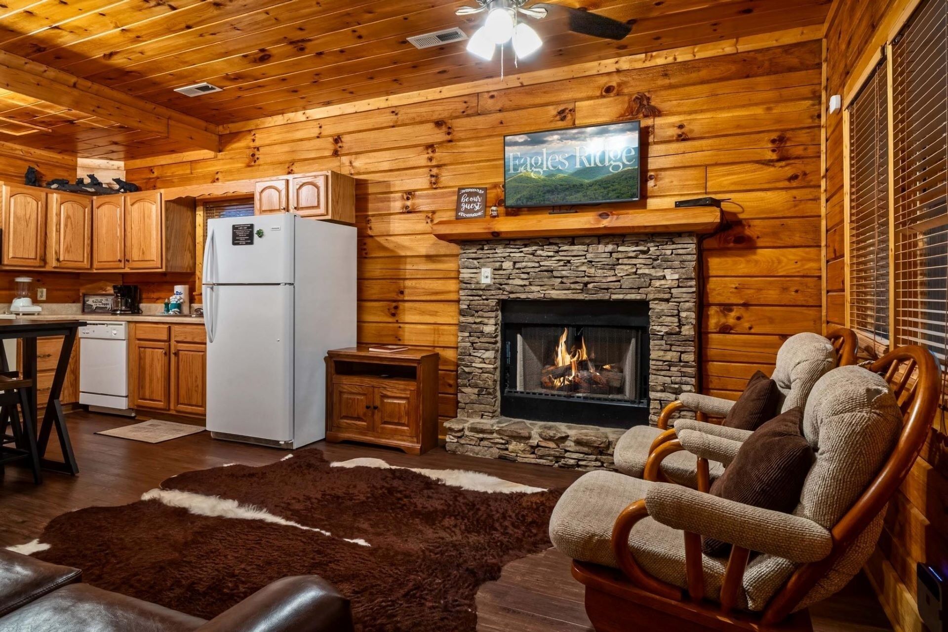 A cozy cabin interior with a stone fireplace, wooden walls, a kitchen area, and a seating area with a fur rug.