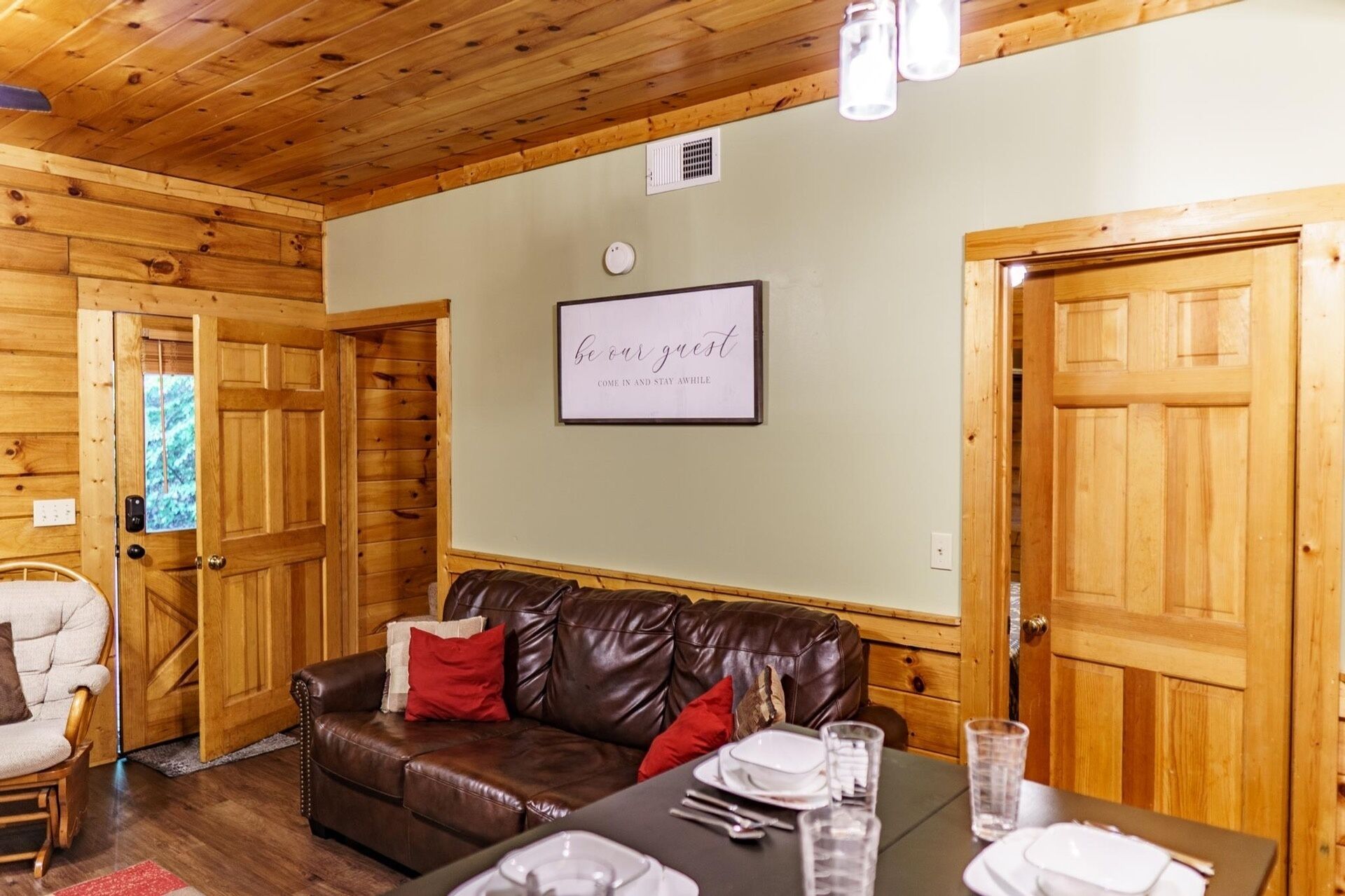 A cozy cabin interior with a leather sofa, a dining table set with plates and glasses, and wooden doors.