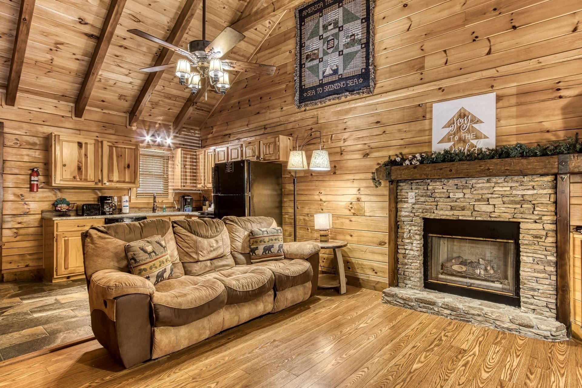 A cozy cabin interior with a wooden ceiling, a stone fireplace, a sofa with animal-themed cushions, and a kitchen area with wooden cabinets and a refrigerator.