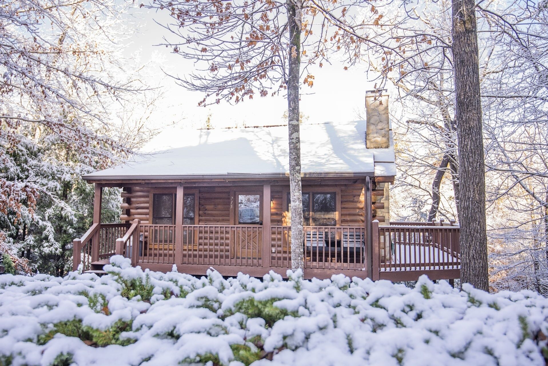 A log cabin with a porch and a chimney, surrounded by snow-covered trees.