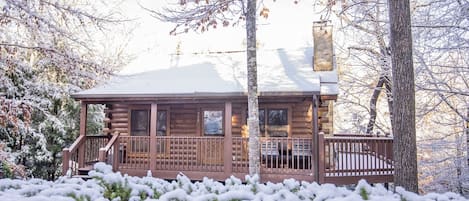 A log cabin with a porch and a chimney, surrounded by snow-covered trees.