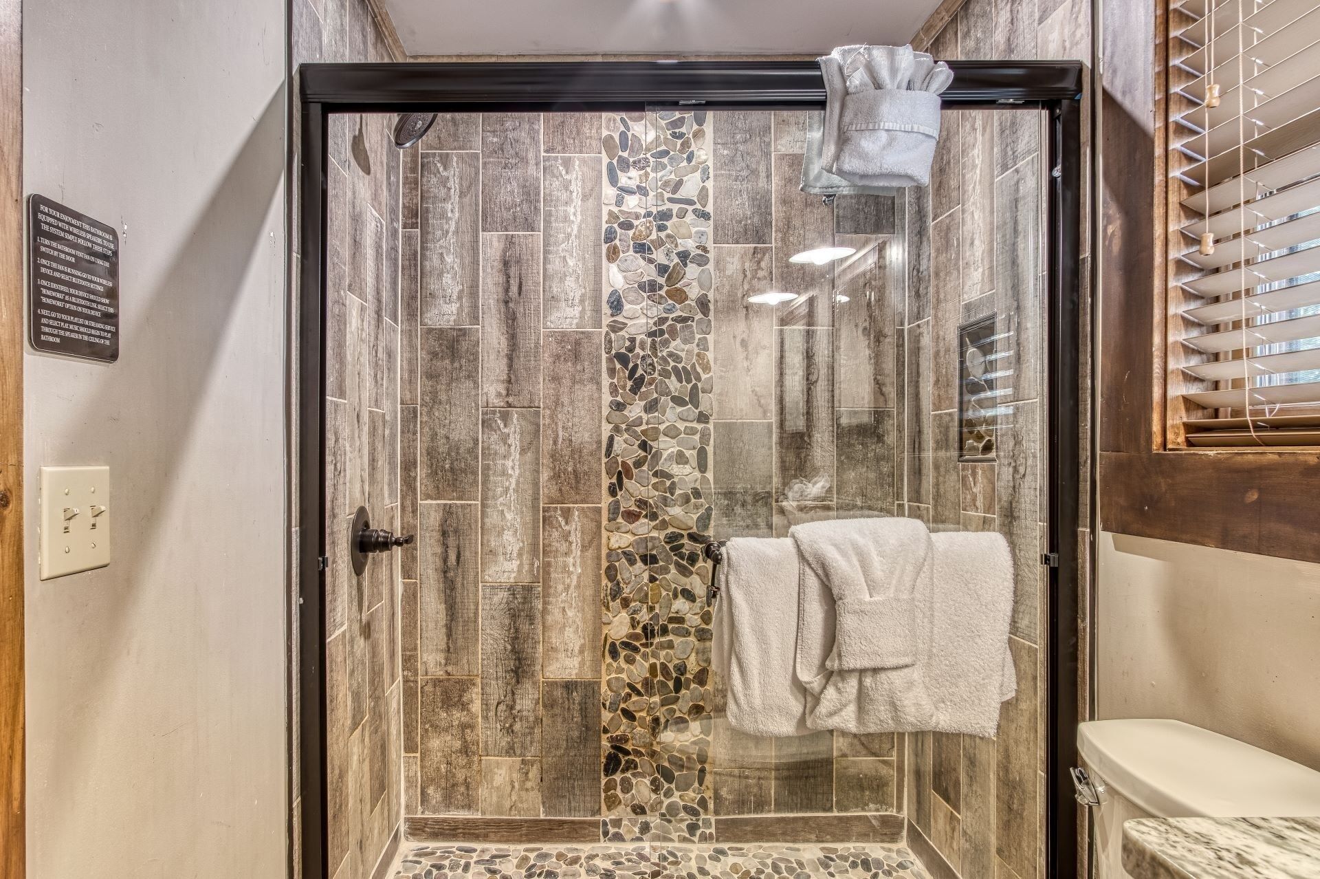 A glass-enclosed shower with a stone mosaic wall, a towel hanging on the door, and a wooden-framed window.