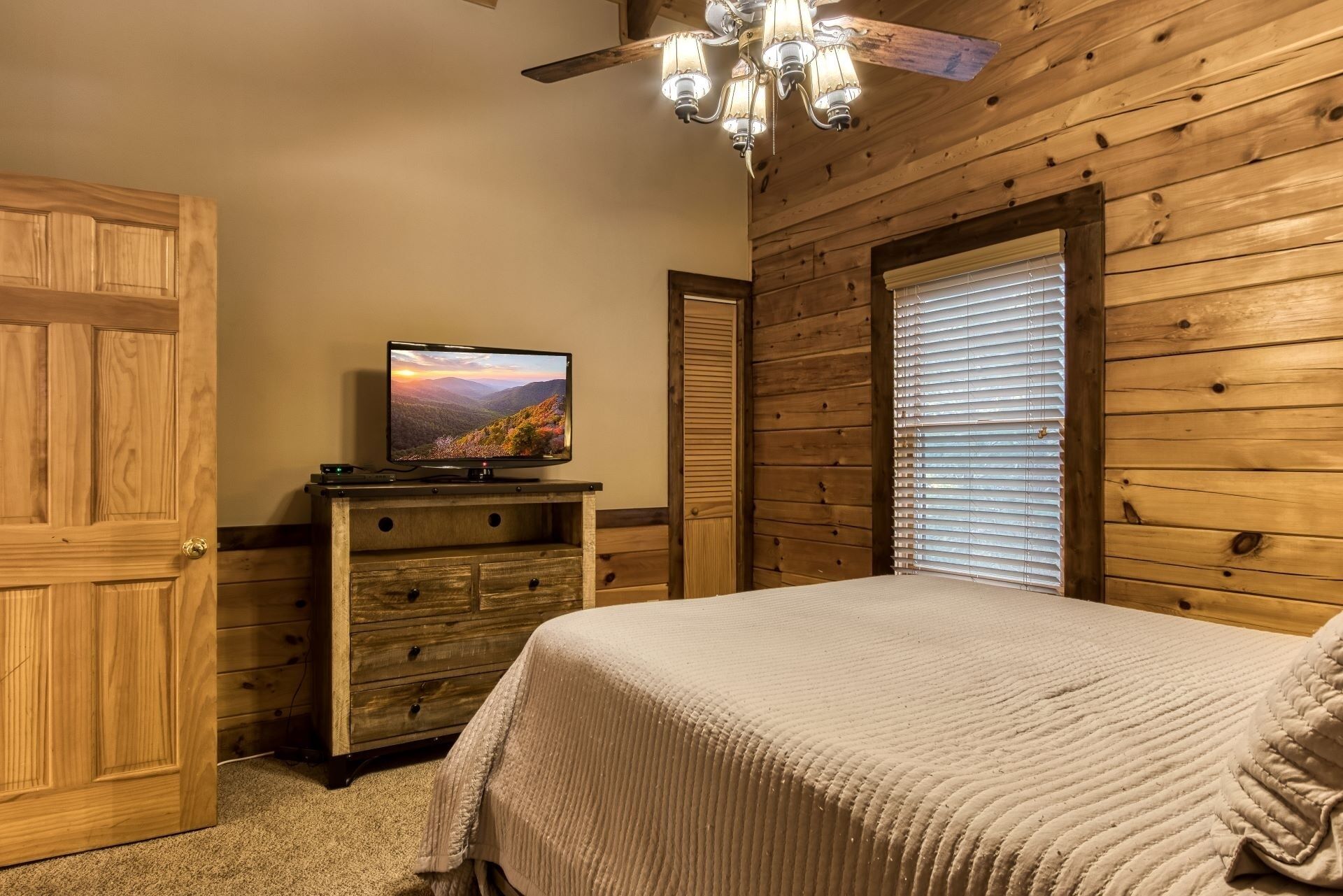 Una camera da letto con soffitto in legno, un letto con coperta beige, una TV su una cassettiera in legno e una finestra con persiane.