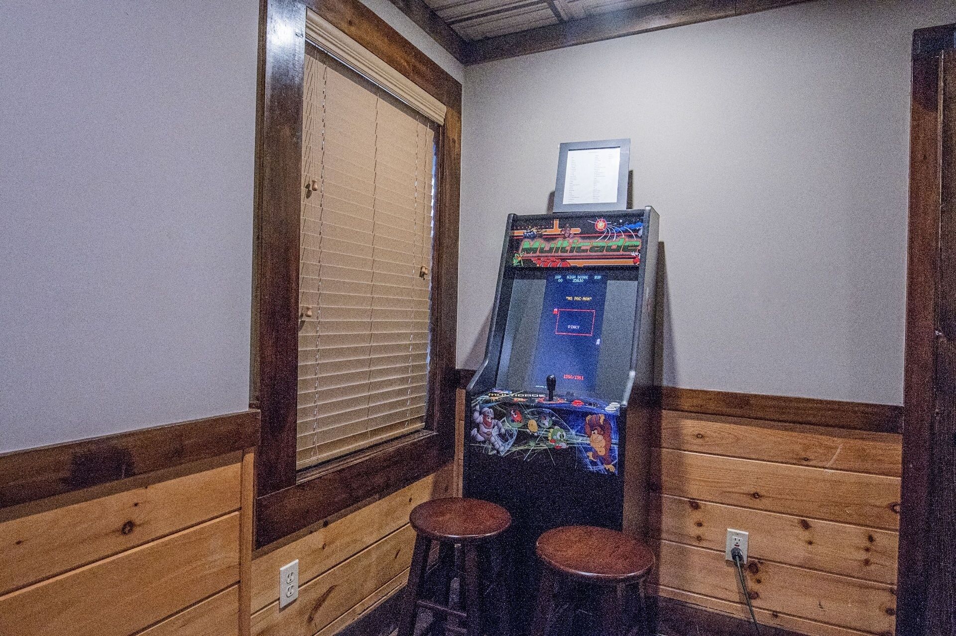Une petite pièce comprenant une seule borne d’arcade, deux tabourets et une fenêtre avec des stores.