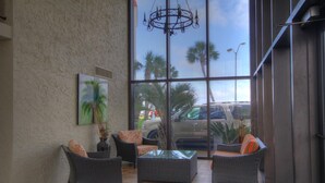 A lobby with wicker furniture, a glass table, a chandelier, and a large window with a view of palm trees and parked cars.