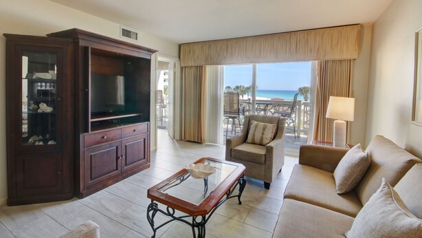 A hotel room with a large window offering a view of the beach, a wooden cabinet, a glass coffee table, and a beige sofa.
