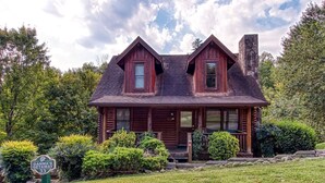 A rustic log cabin with a steeply pitched roof, a stone chimney, and a front porch. - Er33 Ganma's Getaway Great Location, close To Town 4 Bedroom Cabin by RedAwning (Pigeon Forge)