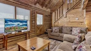 A log cabin interior with a wooden staircase, a sofa, a coffee table, and a television displaying a scenic image. - Er33 Ganma's Getaway Great Location, close To Town 4 Bedroom Cabin by RedAwning (Pigeon Forge)