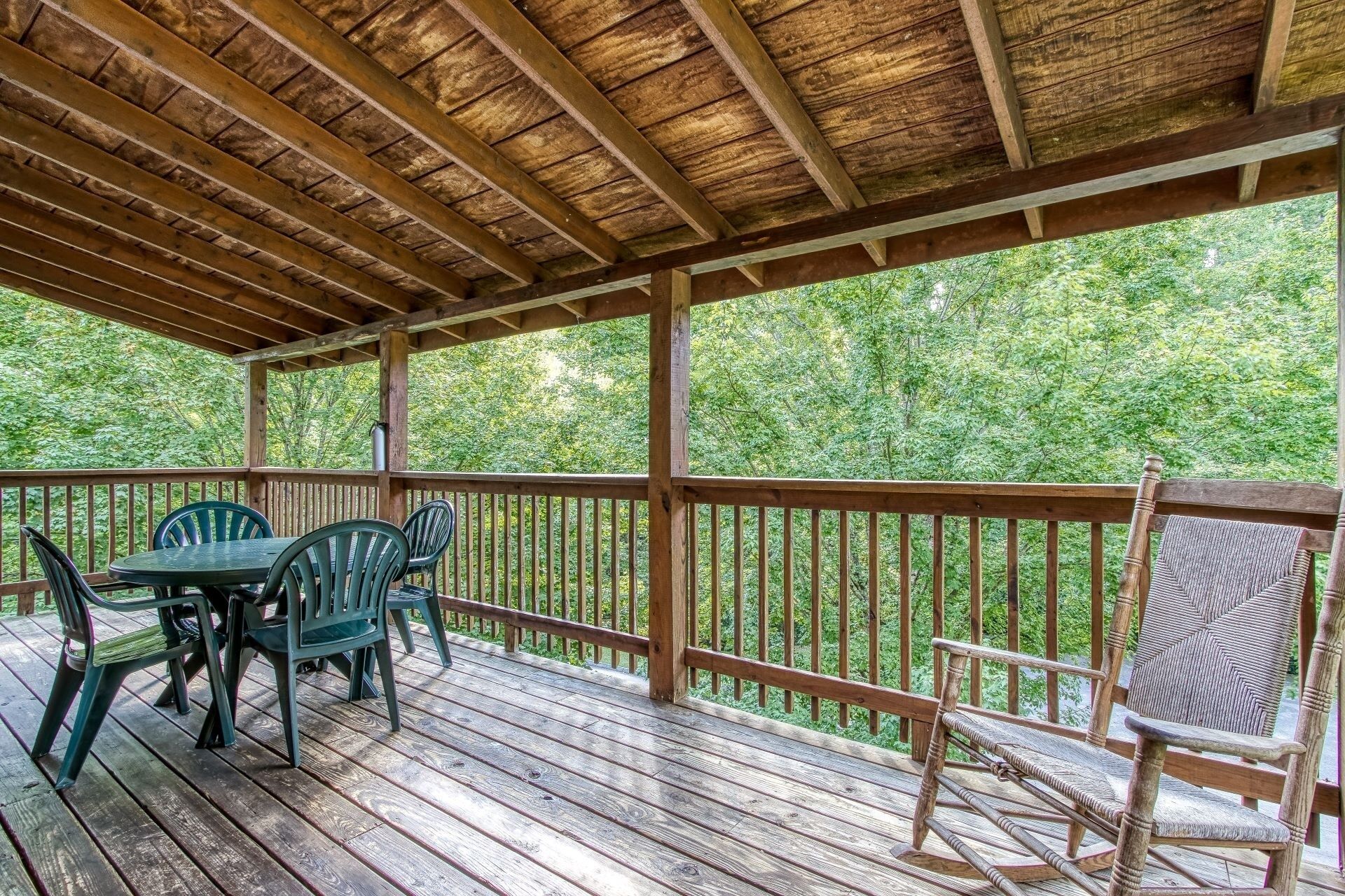 A wooden deck with a table and chairs, surrounded by a wooden railing, overlooking a forested area.